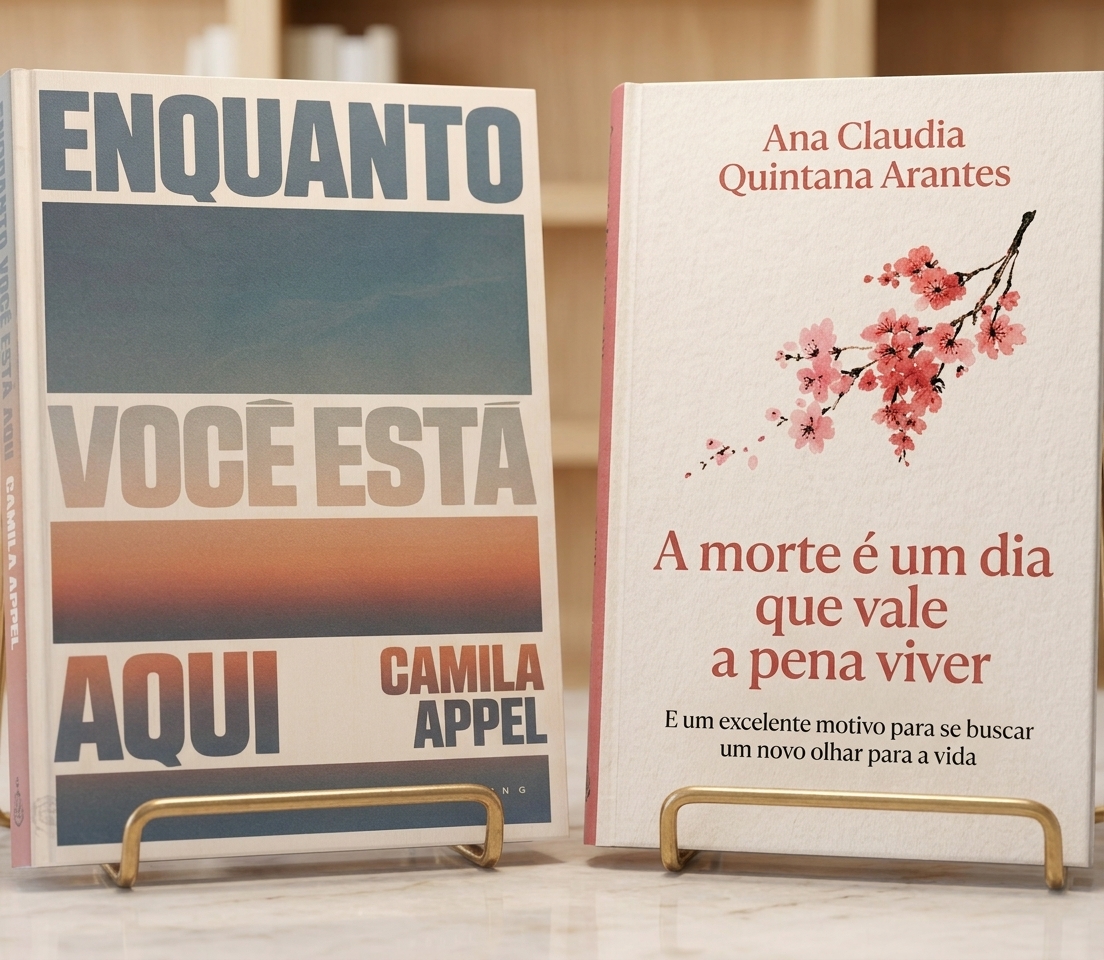 Presença e cuidado: Obras de Ana Claudia Quintana Arantes e Camila Appel.
Para cego ver: Em primeiro plano, sobre uma bancada clara de acabamento refinado, estão dispostos dois livros em suportes metálicos dourados. À esquerda, a capa de Enquanto Você Está Aqui, de Camila Appel, exibe faixas horizontais com tons degradê de azul e alaranjado. À direita, a capa de A Morte É um Dia Que Vale a Pena Viver, de Ana Claudia Quintana Arantes, apresenta fundo branco e o desenho delicado de um ramo com pequenas flores cor-de-rosa. O fundo da imagem traz prateleiras de madeira clara levemente desfocadas.
