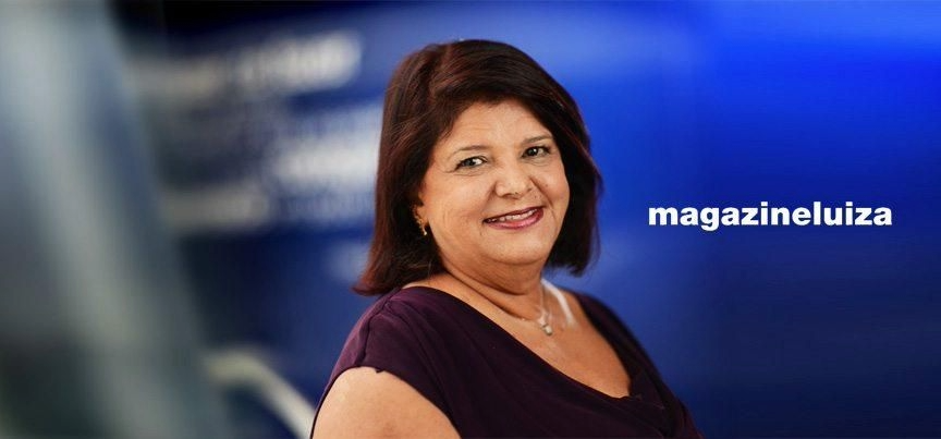 Retrato de Luiza Trajano sorrindo levemente em um ambiente corporativo com iluminação natural, tendo o logotipo da Magazine Luiza, empresa que ela foi gestora por mais de 20 anos e visível ao fundo.