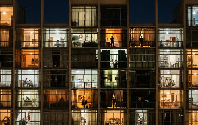 Fotografia noturna de uma fachada de apartamentos do edifício Copan, em São Paulo. As janelas iluminadas mostram silhuetas de moradores em atividades cotidianas dentro de suas casas.